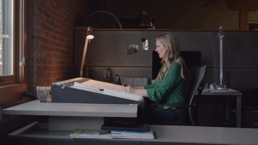 This video features a woman with long blonde hair dressed in a green shirt intently working at an adjustable drafting table she appears to be sketching or drawing with a bright desk lamp illuminating her work surface in a welllit office or studio se