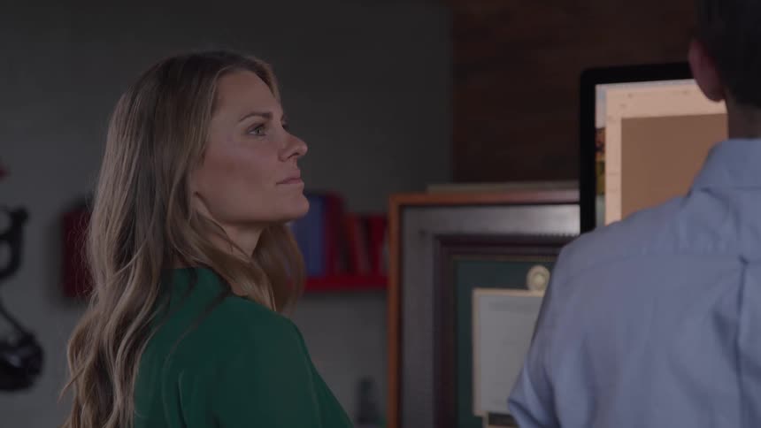 This video shows a blondehaired woman in a green top looking towards a computer screen presumably engaged in a discussion or collaborative work with a person whose back is visible the setting appears to be an office with an award or diploma on the wall