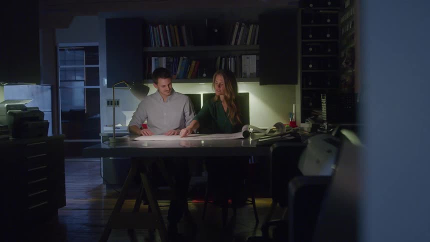 Two colleagues work late in a softly lit office reviewing plans spread across a large desk