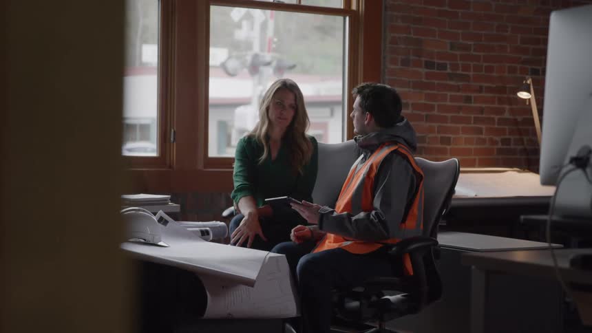 A woman and a man in a safety vest sit by a window in a brickwalled office engaged in a focused discussion