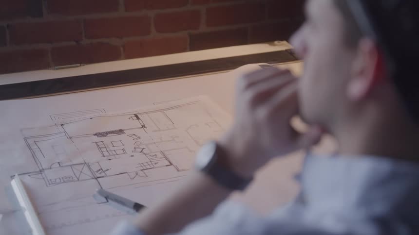 A focused male architect studies a floor plan sketch at a drafting table deep in thought with his hand near his face