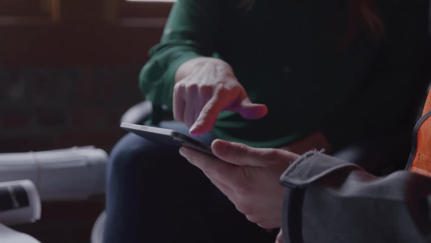 Two people one wearing a green shirt and the other an orange vest interact with a tablet with the person in green using their finger to tap on the screen held by the other