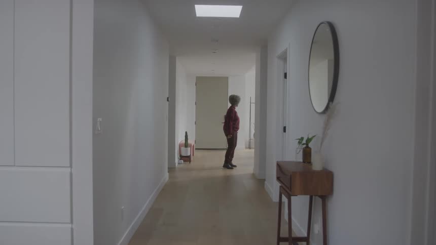 An elderly woman stands at the end of a brightly lit hallway dancing with her arms raised in a modern home