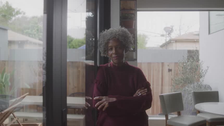 Older woman smiles while leaning against a glass door at home