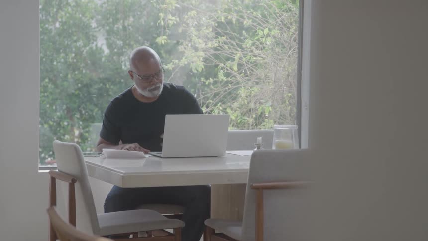 The man is using his laptop and writing on a piece of paper on the dining room table