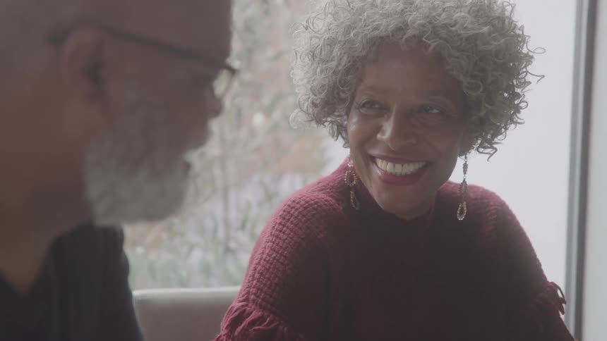 An elderly couple sits in front of a window smiling at each other