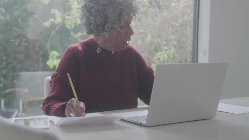 A black woman works on a document at the dining table with a laptop open in front of her at midday