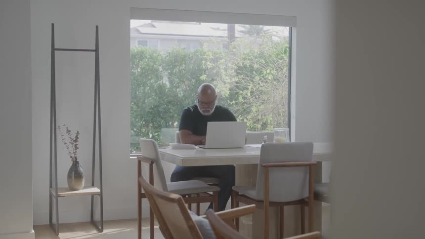 An elderly man sits at a kitchen table typing on a laptop computer