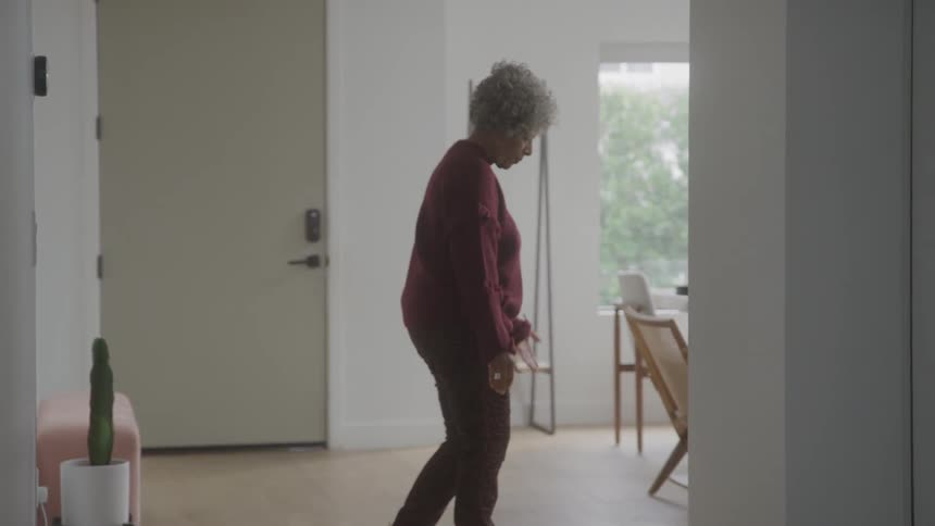 Woman walks slowly through a sparse softly lit room with neutral decor and a cactus in the corner