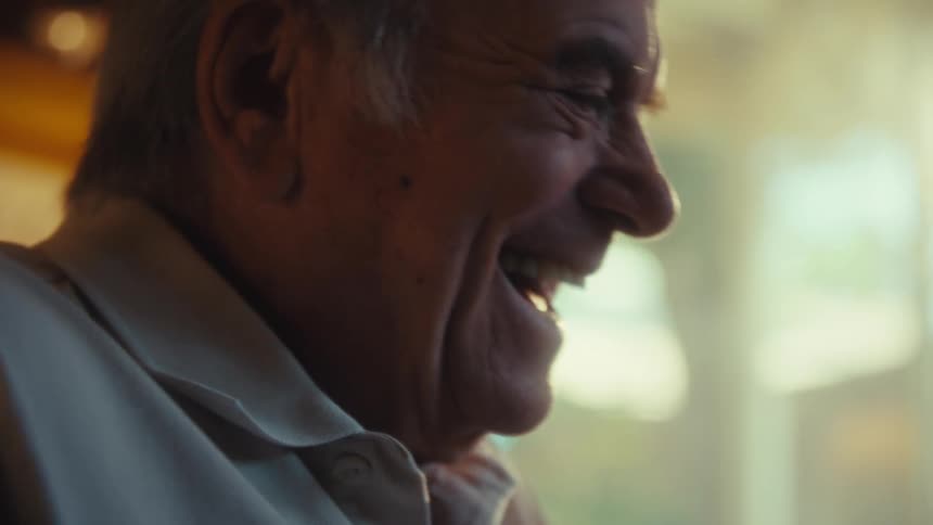 An elderly man smiles warmly in soft natural light while laughing on a phone in his hands