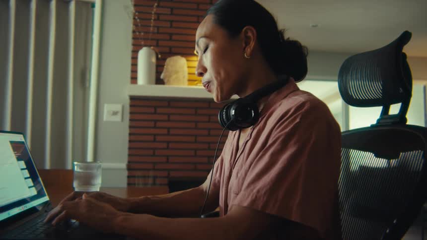 A woman wearing headphones around her neck sits at a table intently typing on a laptop with a glass of water and documents nearby and a brick fireplace visible in the background