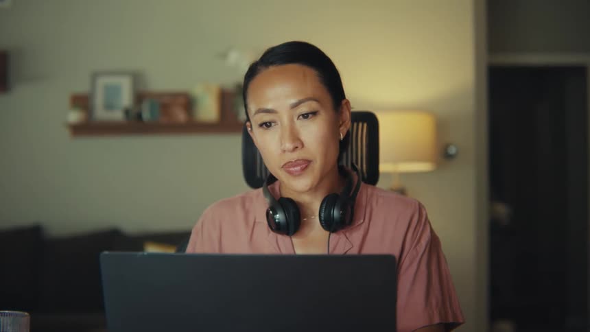 A young woman sits at a desk in her living room wearing a headset draped over her shoulder working on a laptop a glass of water is placed beside the laptop and a couch is behind her with a lamp positioned to the right