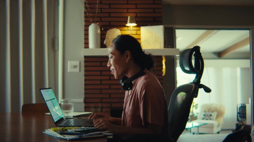 A woman with long dark hair wearing a casual top and headphones around her neck is seated at a wooden desk in what appears to be a home office she is focused on a laptop screen occasionally looking up and smiling