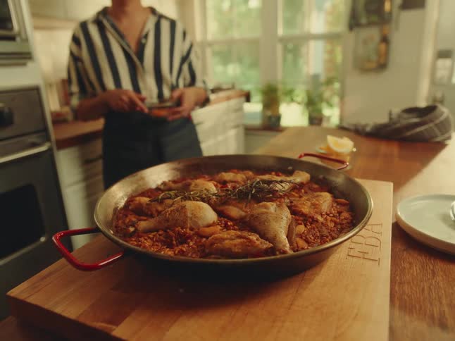 A closeup shot features a large paella pan filled with chicken and rice garnished with herbs sitting on a wooden cutting board in the background a person in a striped shirt and apron is visible in a bright kitchen seemingly preparing more ingredient