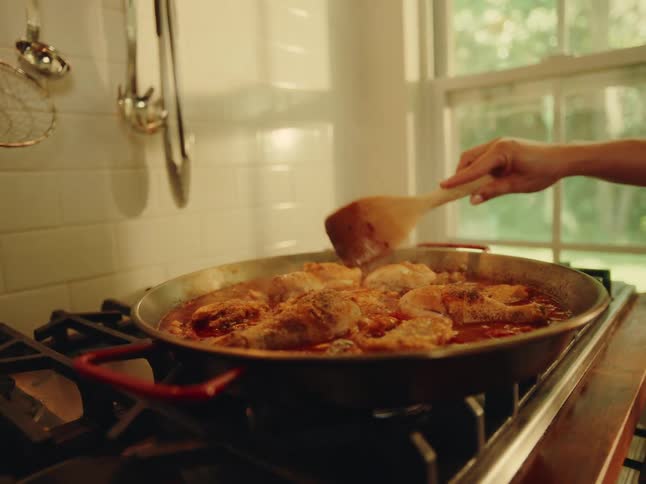 Woman drizzles liquid onto a chicken dish cooking on the stove top at home during the daytime