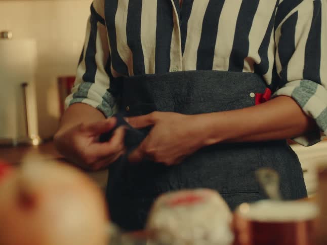 A closeup shot focuses on a person wearing a striped shirt and a dark apron tying the aprons strings around their waist the foreground is blurred showing what appears to be fresh produce on a counter suggesting a cooking or food preparation setting