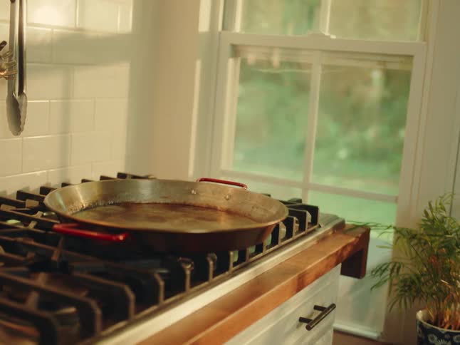 A paella pan rests on a stovetop in a welllit kitchen sunlight streams through a large window illuminating the white tiled backsplash and the wooden countertop