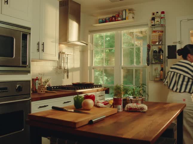 A young adult woman with her hair in a bun wearing a striped shirt stands in a brightly lit kitchen preparing vegetables and taking raw chicken from a tray to a cutting board with various cooking ingredients and utensils on the counters and a window l
