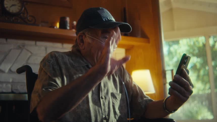 An elderly man wearing a cap and oxygen tubes is seated in a wheelchair happily engaging in a video call on his smartphone gesturing and smiling in a sunlit living room
