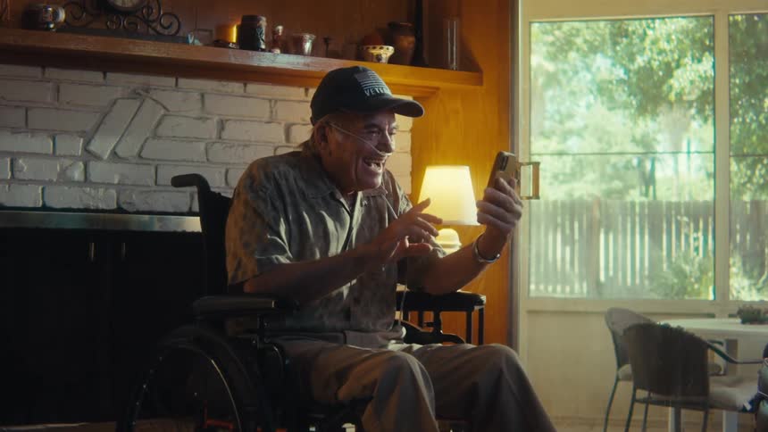 The video shows a man in a wheelchair happily interacting with a smartphone seemingly on a video call he is sitting in a living room with a fireplace and large windows