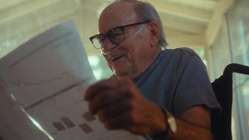 An elderly man sits in a wheelchair reading documents in his hand and smiling with joy wearing reading glasses and a nasal bridle on his nose