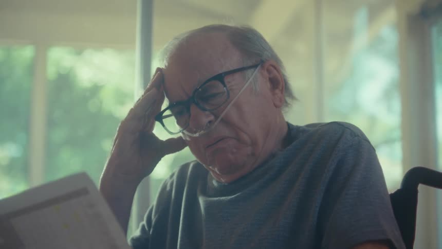 An elderly man sits in a wheelchair beside a glass window reading a document that causes him to frown in sadness a nasal bridle attached to his nose