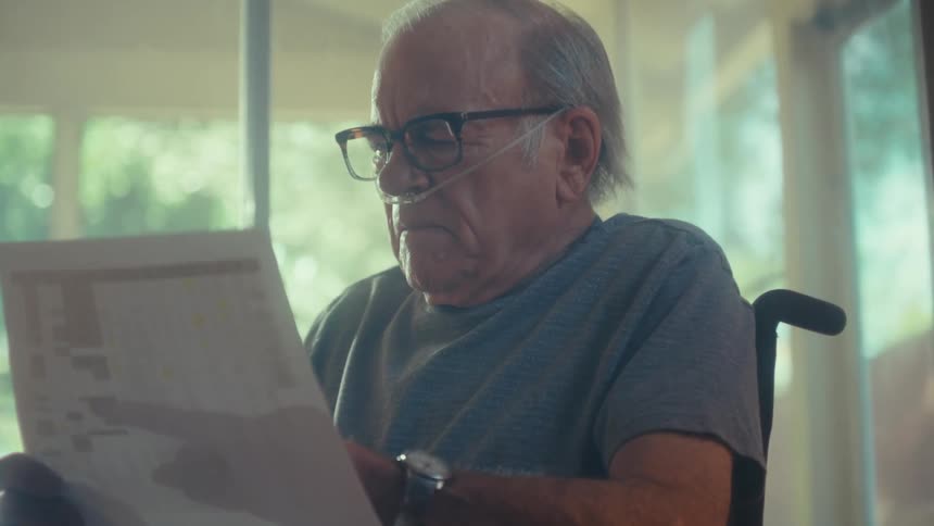 An elderly man in a wheelchair reads a document intently in a warmly lit room