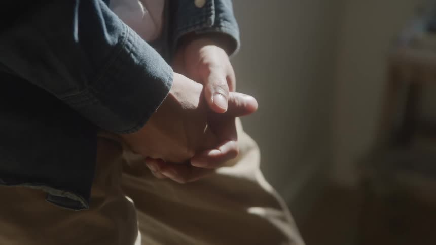 A man stands indoors resting against a wall at midday wearing brown trousers and a denim jacket with his hands clasped below his waist