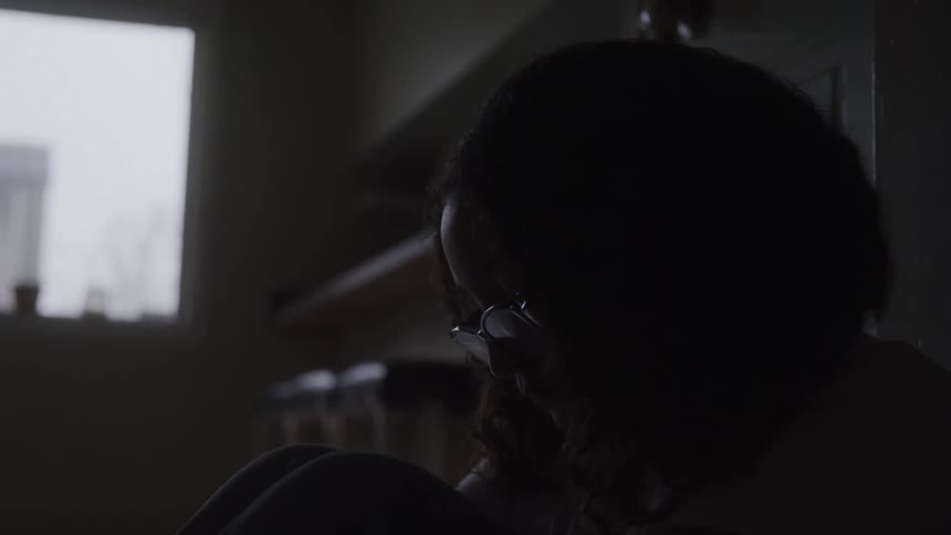 A teenage girl sits indoors in low light looking sad and disturbed