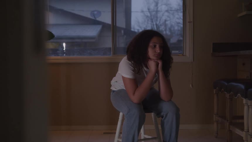 A teenage girl with curly hair sits on a stool indoors looking sad and deep in thought