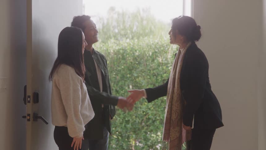 A woman in black clothes is shaking hands with a couple in front of her and they are smiling while standing next to a door