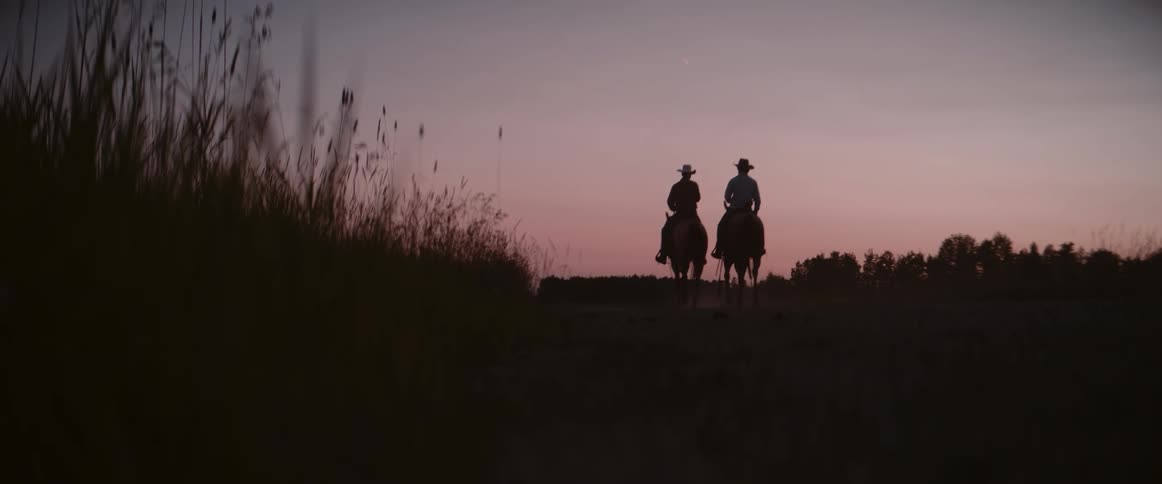 The video captures a serene sunset over a field with tall grasses silhouetted in the foreground two figures on horseback slowly ride away into the fading light disappearing into the distance