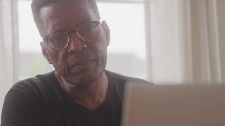 A middleaged man with short dark hair and glasses is seated at a table intensely focused on a laptop screen he then looks up and directly at the camera with a serious expression