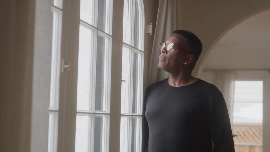 Older man leans against an interior wall while staring out a window