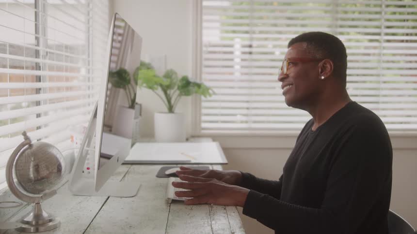 A man with short dark hair and orangeframed glasses is seated at a white wooden desk actively typing on a keyboard while looking at a computer monitor a small globe and a green plant in a white pot are visible on the desk with white blinds covering a