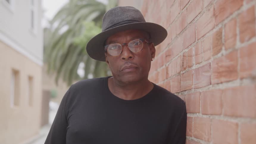 A middleaged man wearing clear glasses a sun hat and a black tshirt rests against a brick wall outdoors at midday