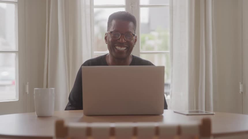 A man with glasses and a black shirt is sitting at a wooden table smiling and waving at a laptop in front of him a white mug and a tablet are also on the table with large windows and white curtains in the background