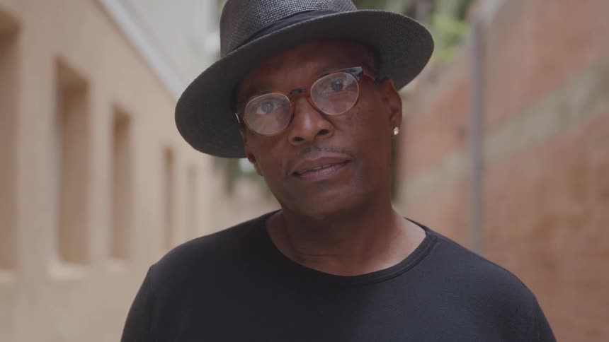 A man wearing a black shirt a grey fedora hat and glasses is looking directly at the camera with a neutral expression he appears to be outdoors with blurred buildings or walls in the background