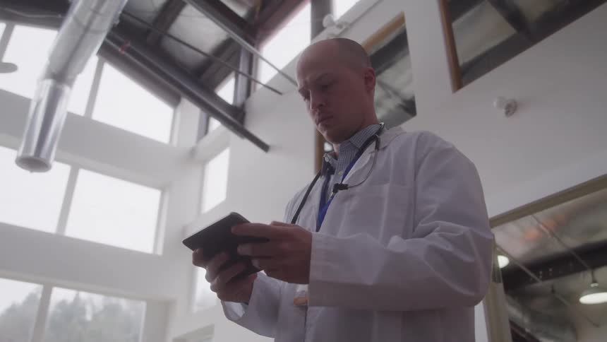 A doctor wearing a stethoscope over his shoulder and a white lab coat stands indoors in an open area at midday using his phone