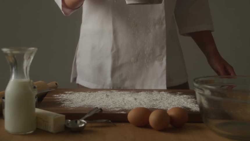 A chef pours flour onto a wooden board in preparation for molding dough with three eggs a milk jar and a bowl in front of them