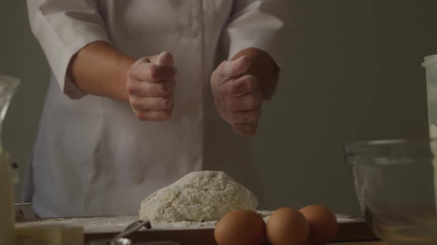 A person wearing a white chefs coat is sprinkling flour from their hands onto a ball of dough on a wooden surface three brown eggs are visible next to the dough and other baking ingredients and tools are blurred in the foreground and background