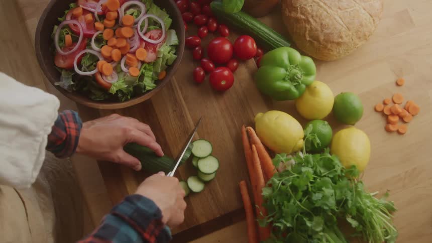 A woman cuts cucumber on a cutting board with a bowl of sliced vegetables on the side and other unsliced vegetables beside the cutting board