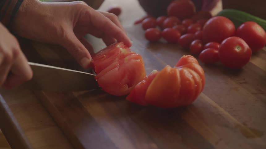 A person is meticulously slicing a large ripe tomato on a wooden cutting board with a sharp knife in the background an assortment of smaller cherry tomatoes and other fresh vegetables are visible indicating a cooking preparation setting