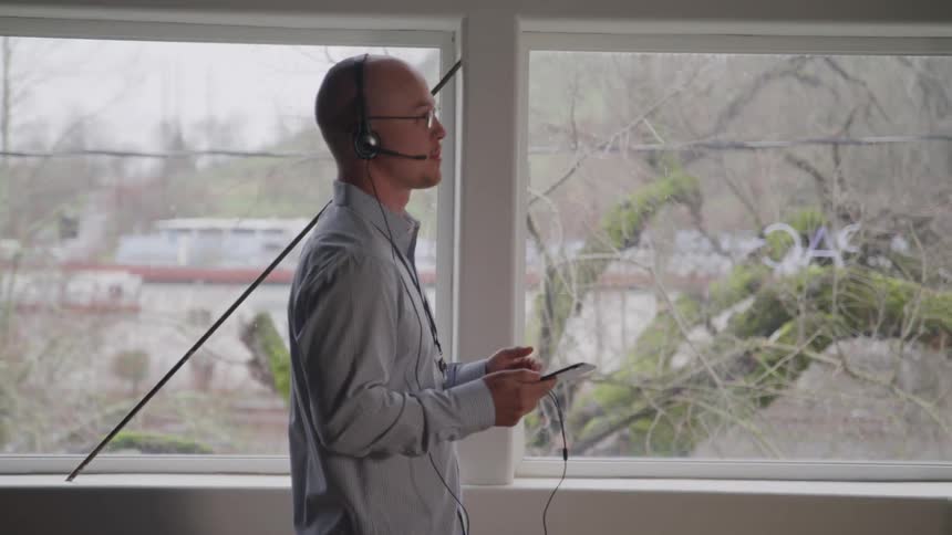 A male professional with glasses and a headset is seen in profile intently interacting with a tablet or phone near a large window overlooking a blurred outdoor scene