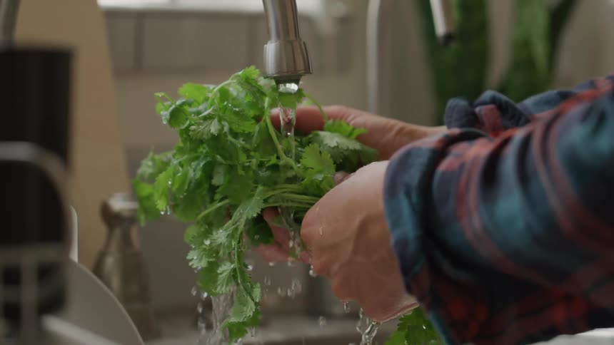 A persons hand holding several green vegetables washing them in water from a tap
