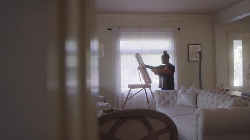 A full shot of a woman engrossed in painting on an easel by a bright window in a comfortably furnished living room