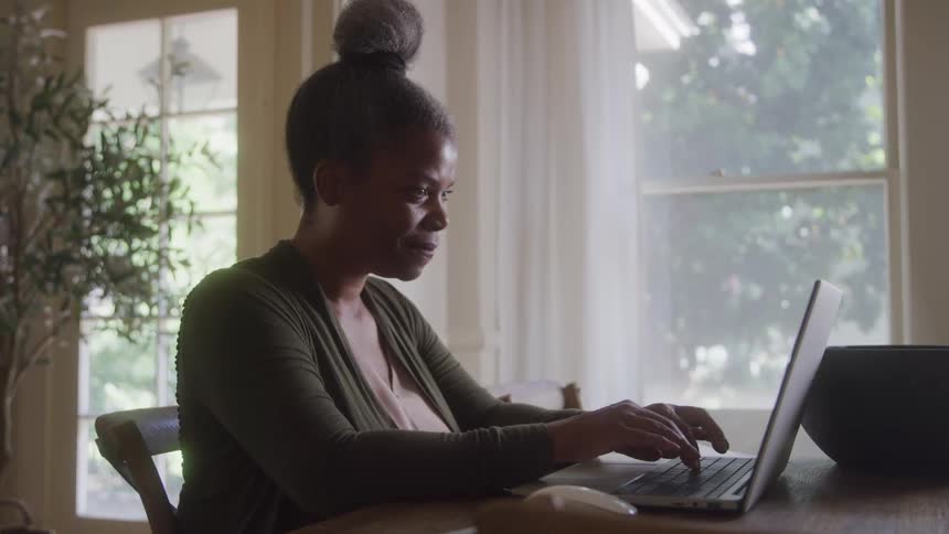 A young woman is shown from a low angle standing in a sunny outdoor setting with trees in the background she is seen typing on a laptop and is wearing her hair in a bun