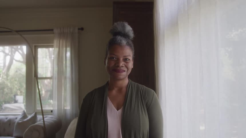 A greyhaired woman with her hair in a high bun stands in an indoor setting looking directly at the camera with a gentle smile the background is blurred showing hints of a living space with a window and curtains