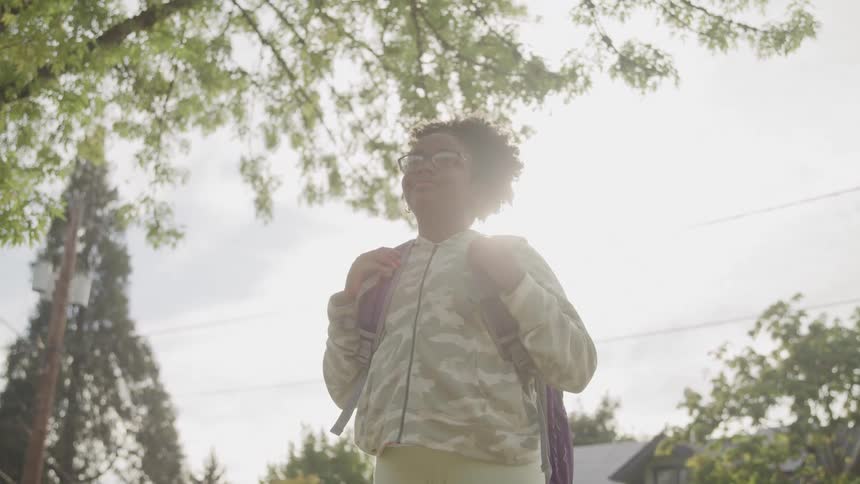 A young girl with curly hair and glasses stands outdoors wearing a camouflage jacket and backpack the shot is taken from a low angle looking up at her against a bright sky with trees in the background