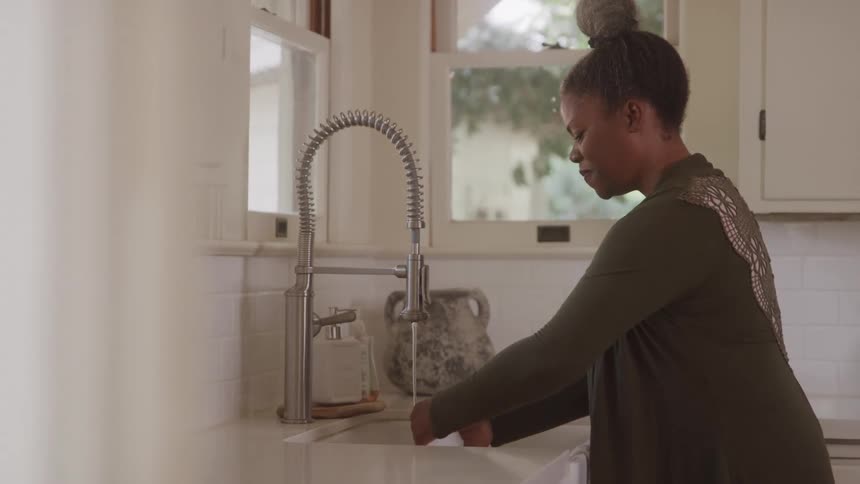 A woman with dark skin and grey hair tied in a bun is standing by a sink washing a white cup under running water she is wearing a green longsleeved shirt